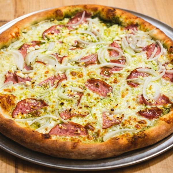 A whole pesto pizza with salami, onions, and feta cheese on a metal pan.
