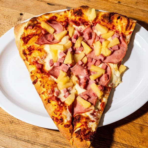 A large slice of Hawaiian pizza with ham and pineapple on a white plate, on a wooden table.